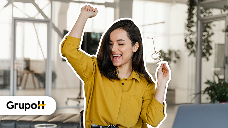 Mulher branca vestida de amarelo, bem feliz em seu ambiente de trabalho, com um óculos em uma das mãos, representando o bem-estar das mulheres