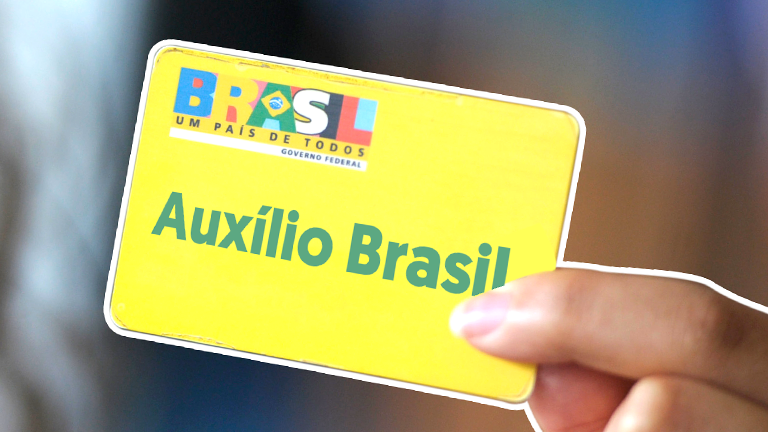 Cartão Auxílio Brasil amarelo sendo segurado por uma mão feminina, representando o benefício liberado pelo governo para pessoas em situação de extrema pobreza e na pobreza